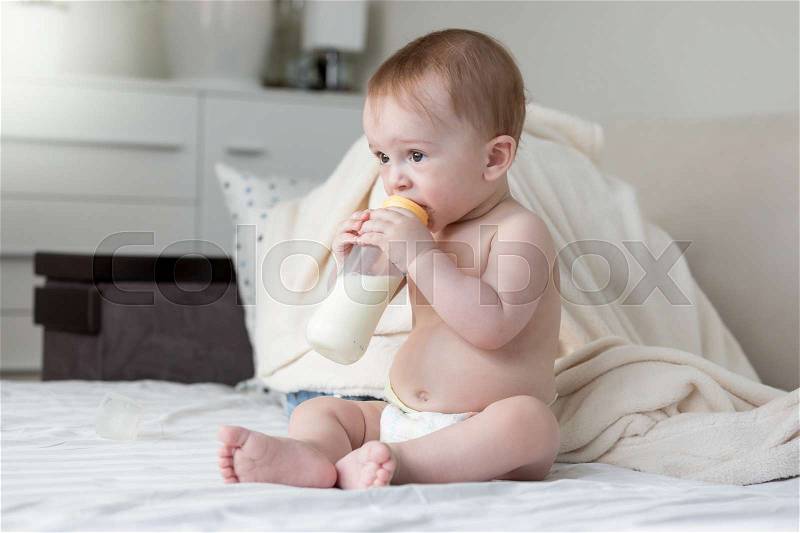 Beautiful 9 months old baby boy sitting on bed and drinking milk, stock photo