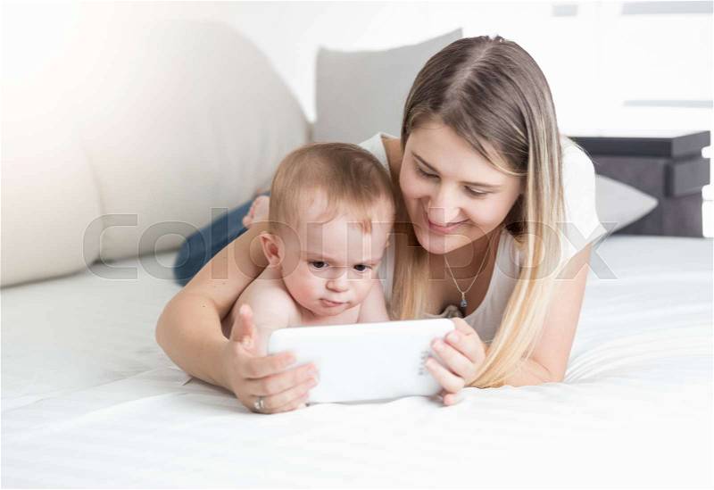 Beautiful young mother lying with her baby on bed and using tablet, stock photo