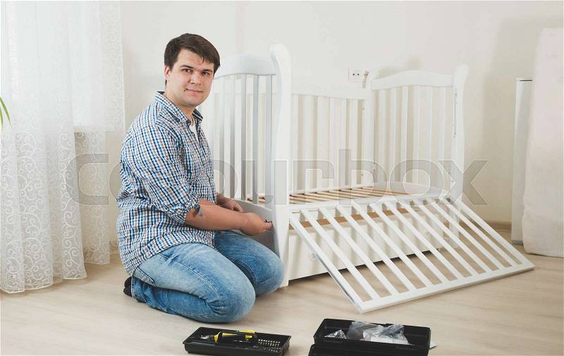 Young man assembling white wooden crib in nursery, stock photo
