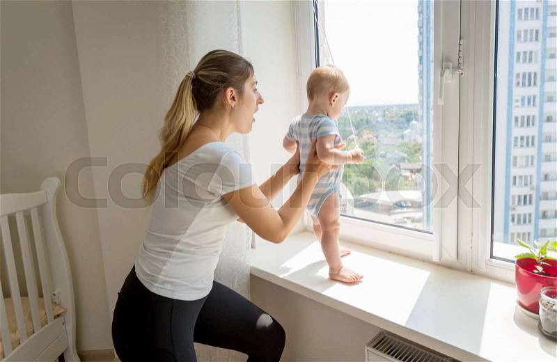 Mother rushing to her baby that is trying to open window, stock photo