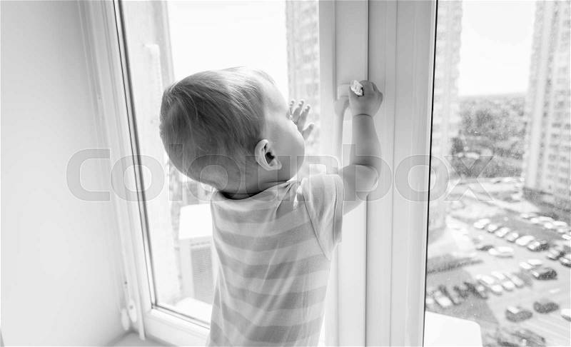Black and white image of baby in danger. Baby boy pulling handle of window, stock photo
