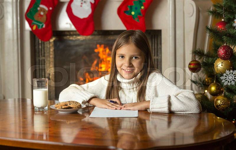 Happy smiling girl sitting by fireplace and writing letter to Santa Claus, stock photo