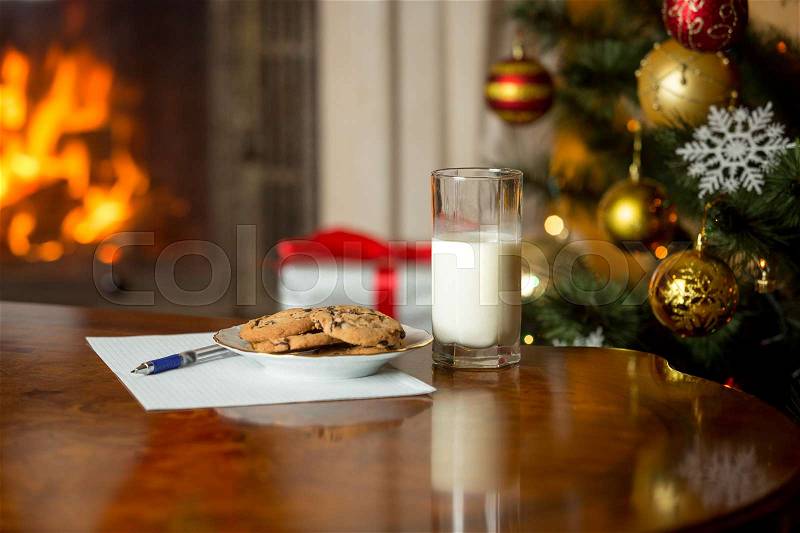 Treats and letter to Santa on wooden table next Christmas tree and burning fireplace, stock photo