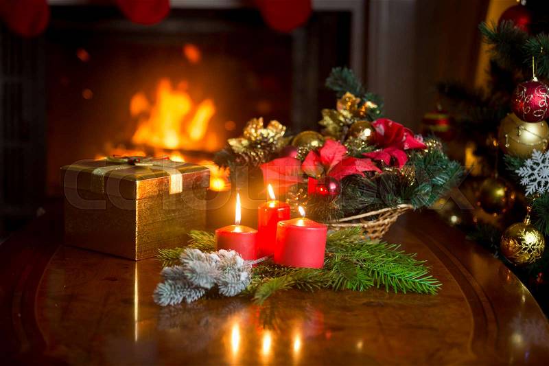 Burning candles, Christmas wreath and golden gift box on table next to burning fireplace at living room. Image with shallow depth of focus, stock photo