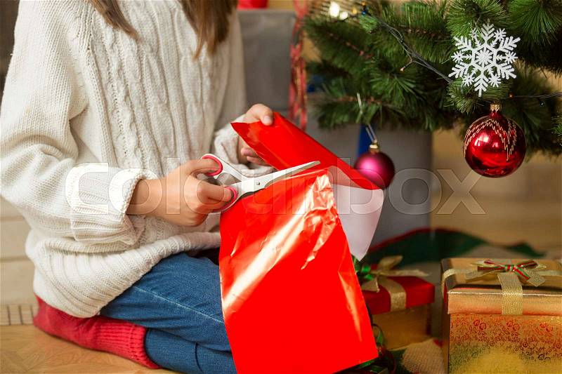 Girl sitting under Christmas tree and cutting red paper with scissors for decorating presents, stock photo