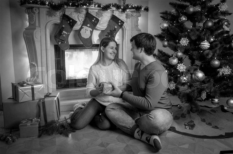Black and white image of handsome young man sitting at fireplace with woman and giving her Christmas present, stock photo