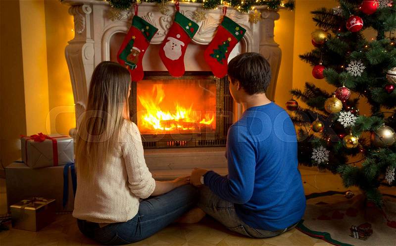 Young couple in love sitting by the fireplace decorated or Christmas and looking at fire, stock photo