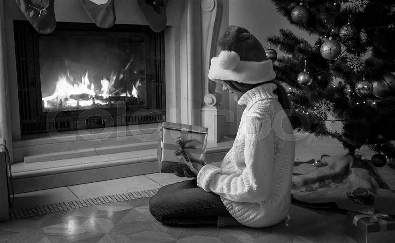 Black and white image of girl holding gift box and sitting next fireplace, stock photo