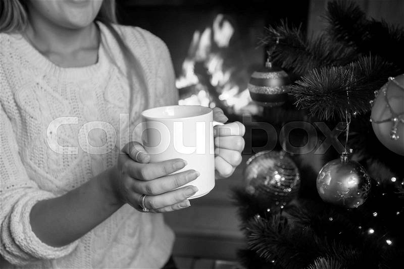 Closeup black and white image of woman sitting at burning fireplace with cup of tea, stock photo