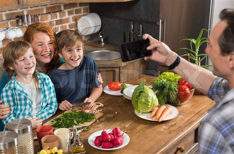 Family In Kitchen Cooking Food Father Taking Photo On Cell Smart Phone Of Mother Son And Daughter Sitting At Table Preparing Healthy Meal, stock photo