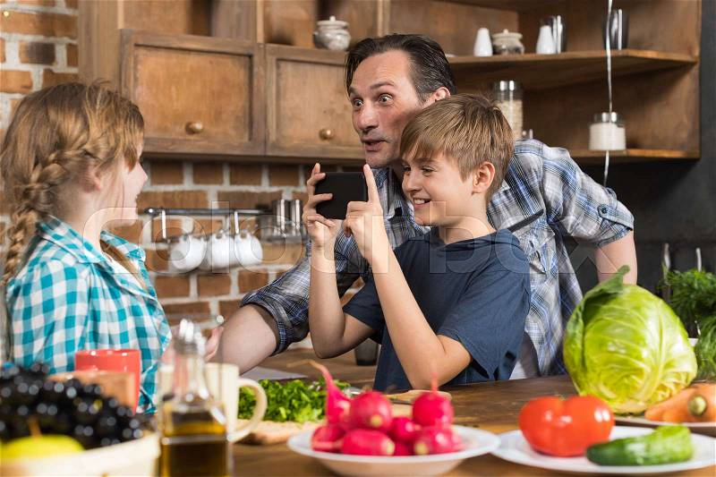 Happy Family In Kitchen, Father And Son Taking Photo Of Daughter Cooking Food On Cell Smart Phone Preparing Healthy Meal, stock photo