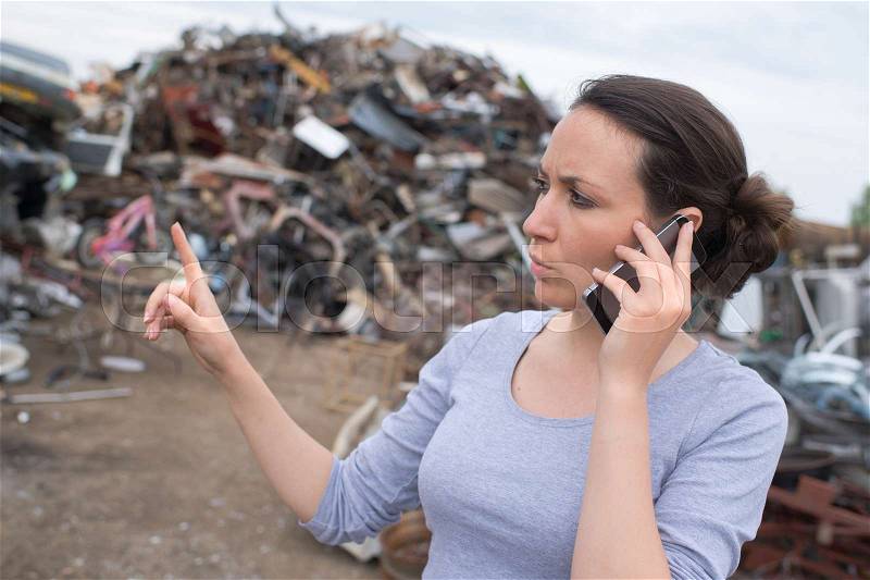 Metal recycling worker gesturing and using mobile phone at junkyard, stock photo