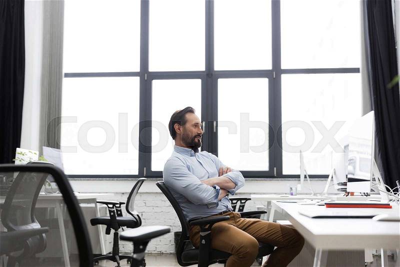 Mature business man sitting on a chair with arms folded and looking at computer screen at the office, stock photo