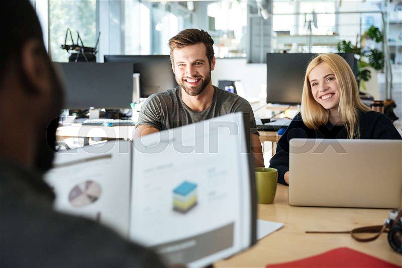Picture of happy young colleagues sitting in office coworking using laptop computers and holding diagrama. Looking aside, stock photo