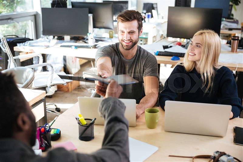 Image of happy young colleagues sitting in office coworking using laptop computers holding documents. Looking aside, stock photo