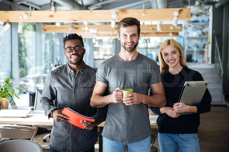 Photo of happy young colleagues in office coworking standing holding laptop, documents and coffee. Looking at camera, stock photo