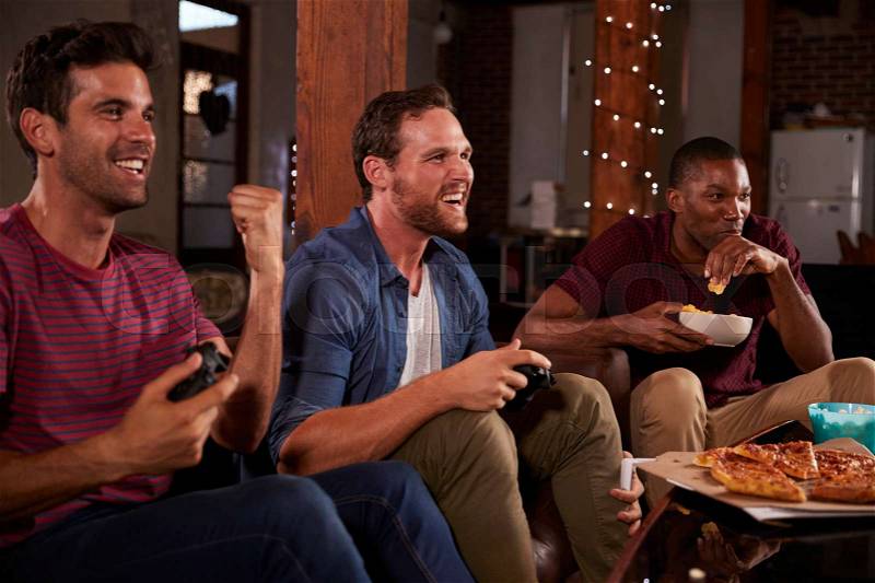 Three male friends playing video games and eating at home, stock photo