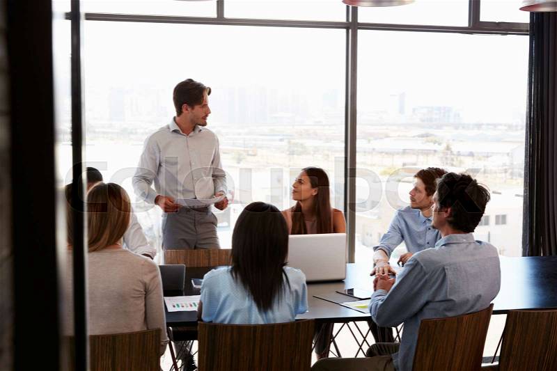 Young man with documents addresses team at boardroom meeting, stock photo