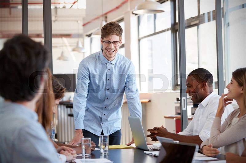 Happy young man stands addressing team at business meeting, stock photo