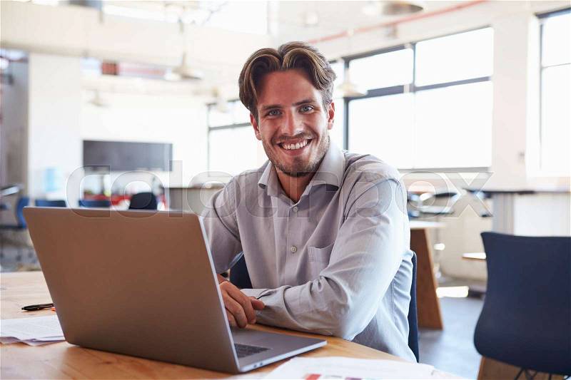 Young man in office using laptop computer smiling to camera, stock photo