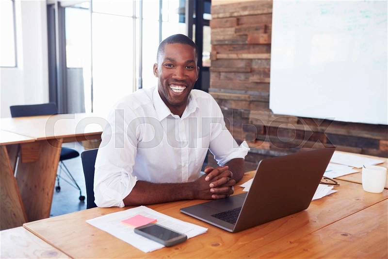 Young black man at desk with laptop computer looks to camera, stock photo