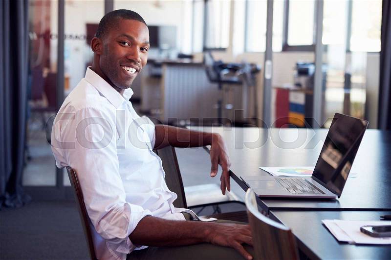 Young black man sitting at desk in office smiling to camera, stock photo