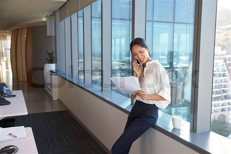 Businesswoman Making Phone Call Standing By Office Window, stock photo