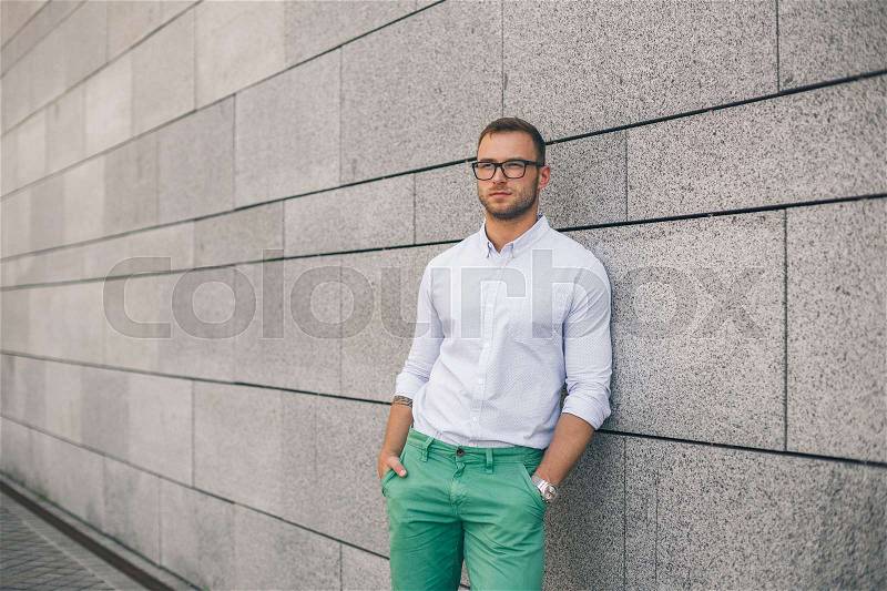 Cheerful young handsome man in sunglasses keeping hands in pockets and looking away with smile while standing against grey background. Stylish concentrated man, stock photo