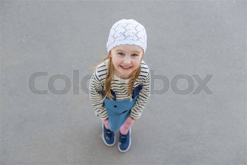 Lovely small girl in white knitted hat standing outside, smiling, wearing striped sweatshirt, stock photo