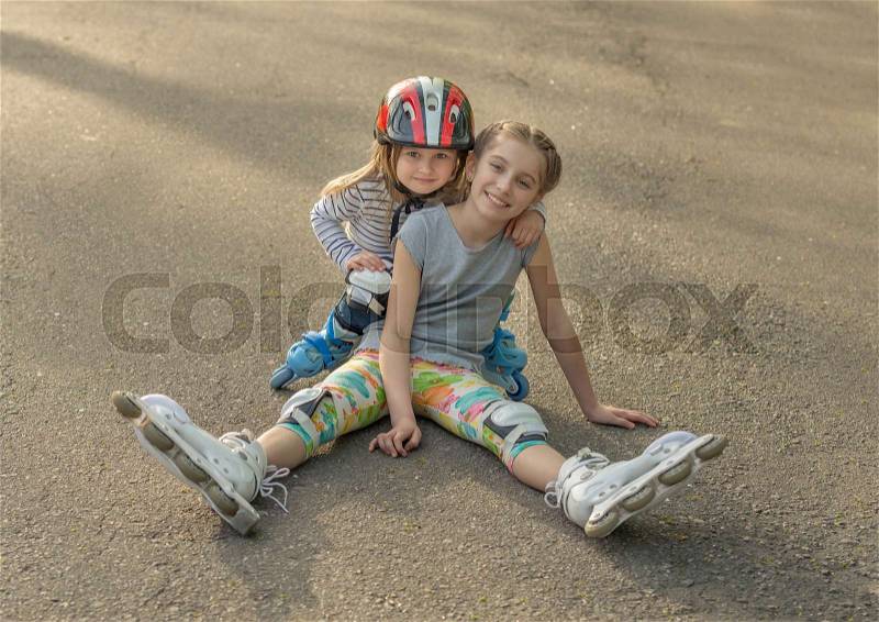 Two lovely sisters roller skating together, wearing protection, resting on the asphalt in park, stock photo