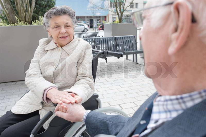 Old couple in wheelchari holding hands outdoors, stock photo
