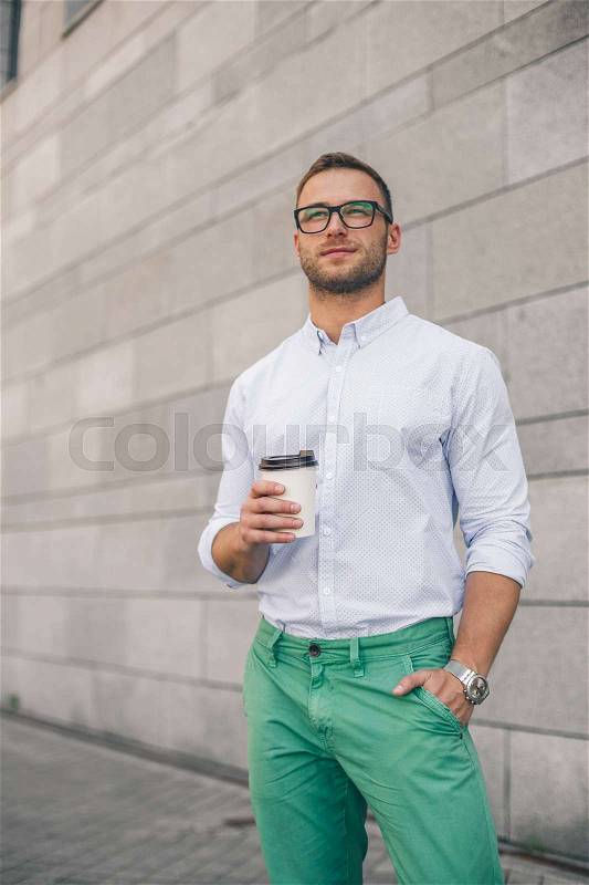 Thoughtful casual man outdoors looking up and smiling. Handsome man in glasses holding coffee cup standing outdoors with cityscape in the background,, stock photo