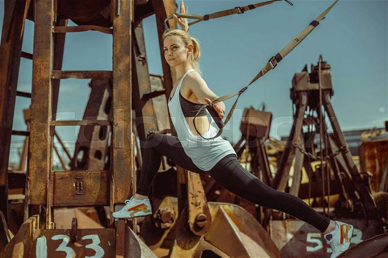 Side view of beautiful young woman doing lunges with trx strips against metal rusty background, stock photo