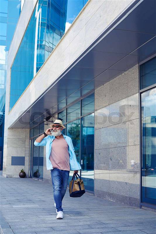 Vertical of fashionable old man talking on phone and walking down street while carrying handbag, stock photo