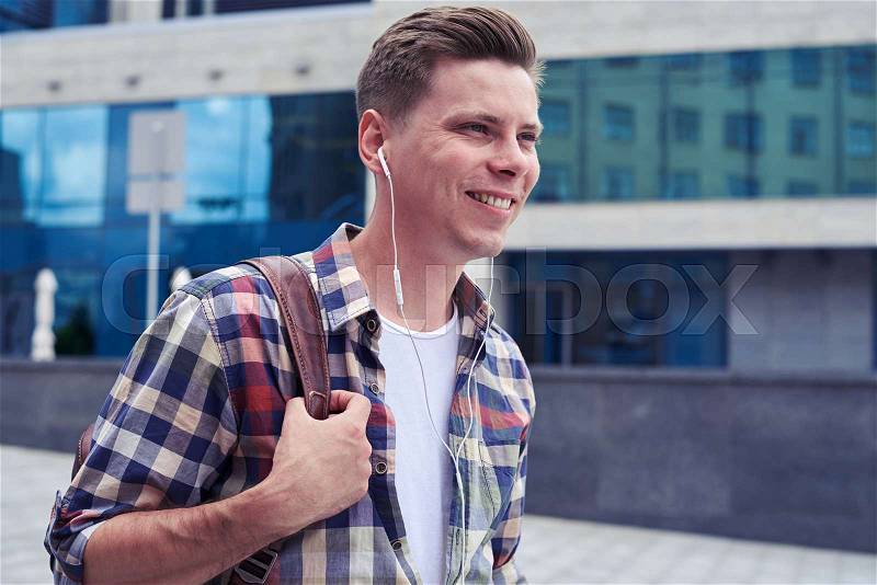 Close-up shot of cute guy listens to music while walking in downtown. Student carrying backpack, stock photo