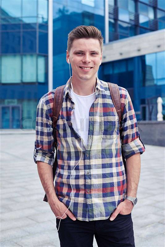 Mid shot of smiling guy in checked shirt with backpack standing near business center. Man listening music , stock photo
