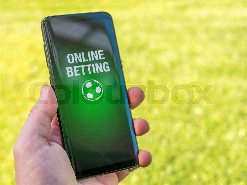 Man holds smart phone with online betting app. Green soccer grass in background, stock photo