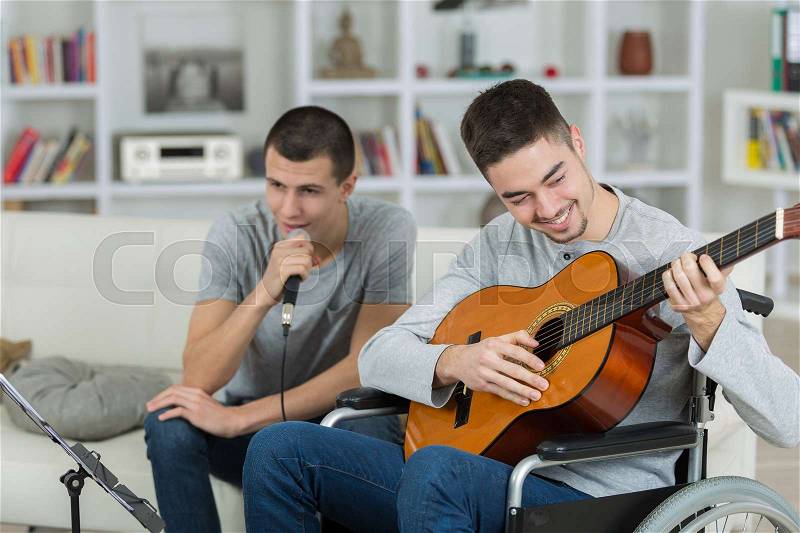 Man in wheelchair playing guitar, friend singing into microphone, stock photo