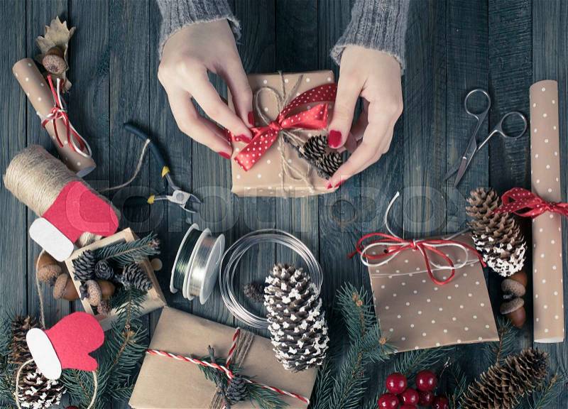Christmas background. Top view of woman hands wrap New Year present. Packed gifts and scrolls, spruce branches and tools on shabby wooden table. Workplace for preparing handmade decorations. Winter holidays concept. , stock photo