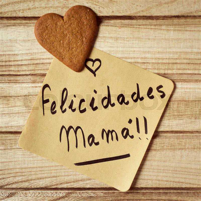 Closeup of a piece of paper with the text felicidades mama, congrats mom written in spanish and a heart-shaped cookie, on a rustic wooden background, stock photo