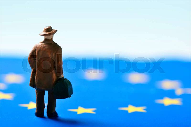 Miniature traveler man seen from behind wearing a hat and carrying a suitcase, on an european union flag, stock photo