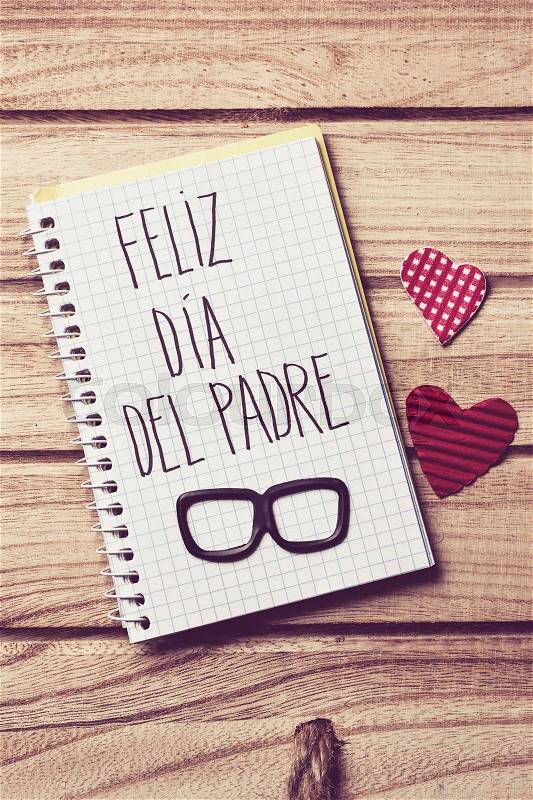 The text feliz dia del padre, happy fathers day in spanish written in the page of a notebook, a pair of black eyeglasses and some red hearts on a rustic wooden background, stock photo