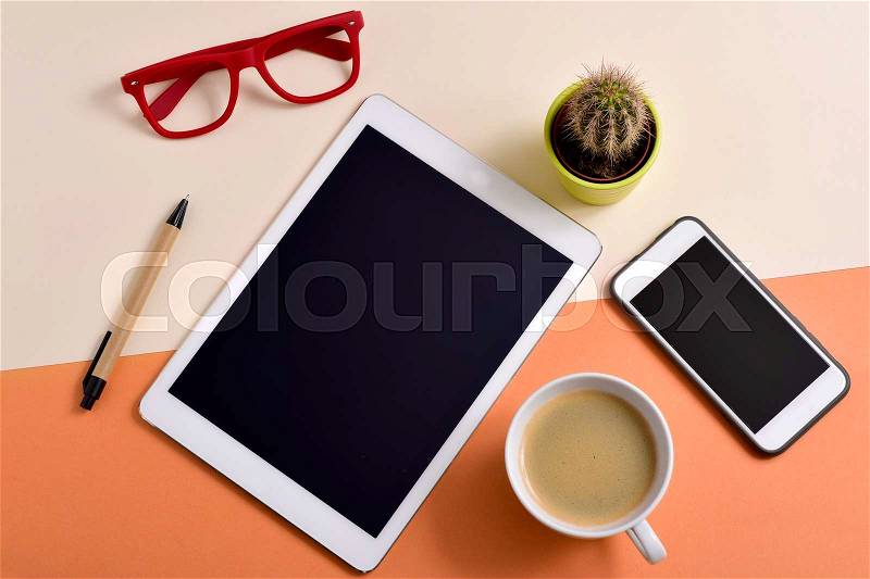 High-angle shot of an office desk full of things, such as a pair of red eyeglasses, a cup with white coffee, a tablet, a smartphone, a pen and a cactus, stock photo