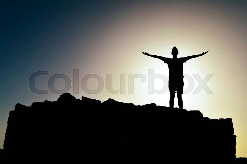 The silhouette of a young caucasian man with open arms in the air, on a monticule, backlighted by the sun at his back, stock photo