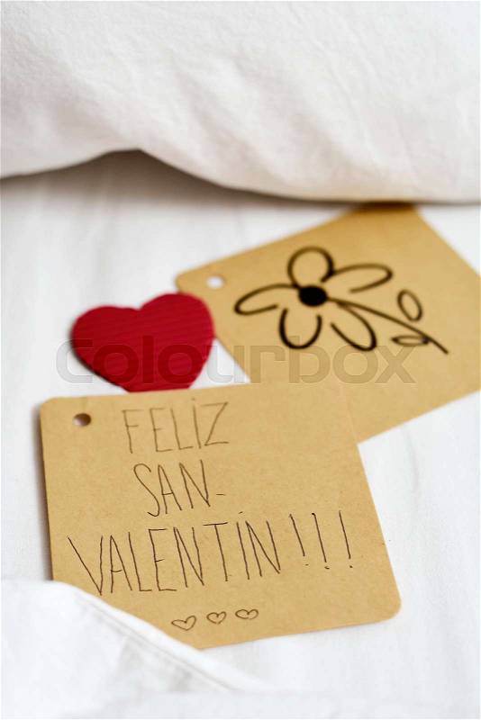 Closeup of a brown paper note with the text feliz san valentin, happy valentines day written in Spanish and a flower drawn in another note, and a red heart, on the white sheets of an undone bed, stock photo