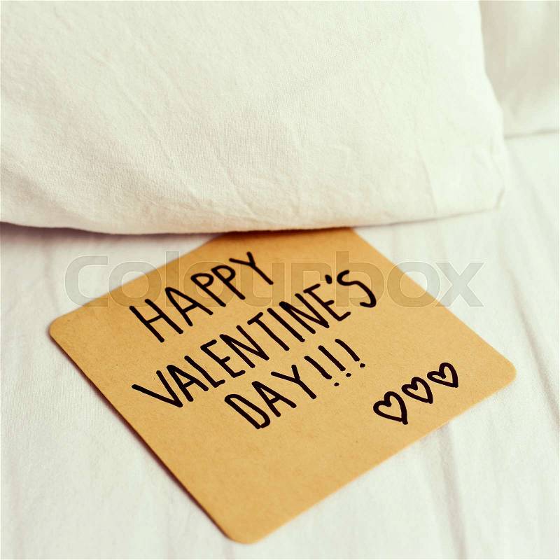 Closeup of a brown paper note with the text happy valentines day written in it, on the white sheets of a bed, stock photo