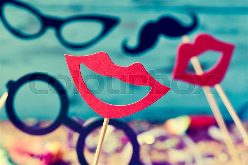 Some different mouths, eyeglasses and mustaches on sticks, against a blue background and a wooden surface full of confetti, stock photo