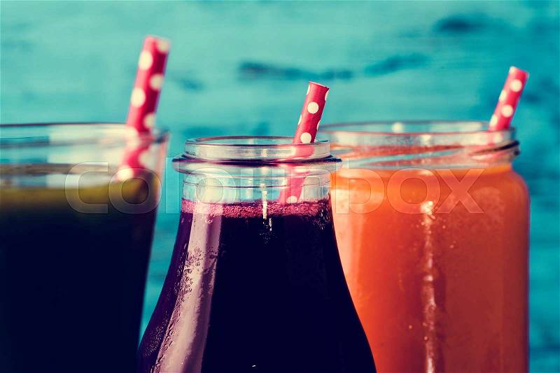 Closeup of three different fresh smoothies served in a glass, a glass jar and a glass bottle with red drinking straws patterned with white dots, against a blue rustic wooden background, stock photo