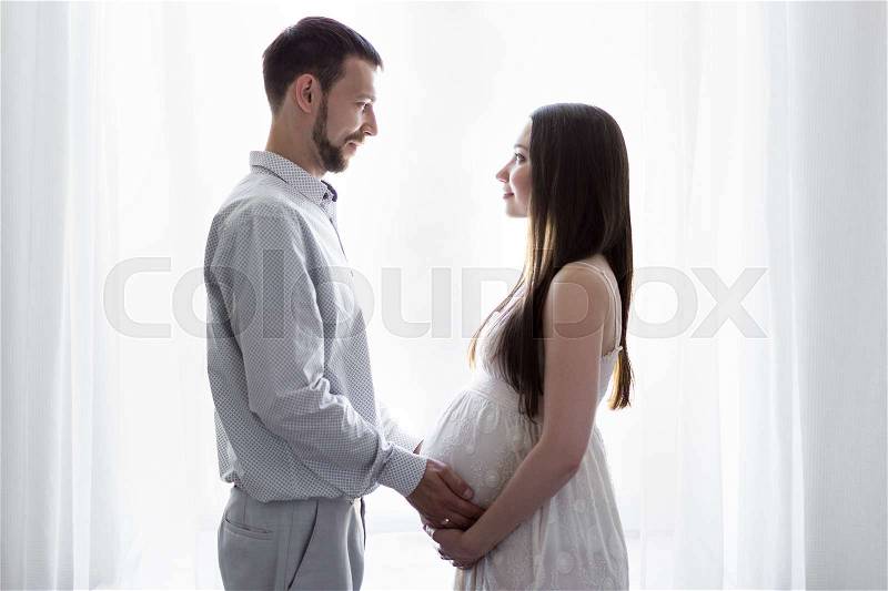 Family, pregnancy and parenthood concept - happy pregnant couple standing in front of the window, stock photo