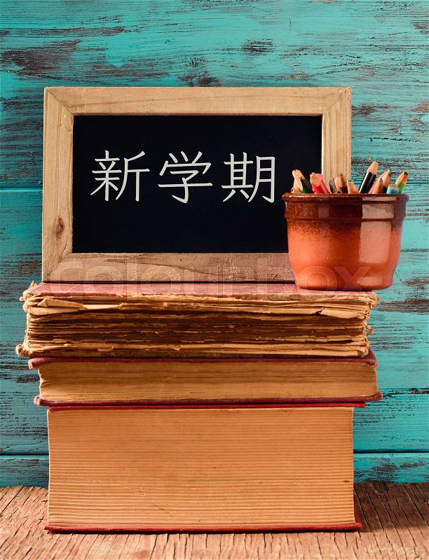 A chalkboard with the text back to school written in Japanese and a pot with some pencils on a pile of old books, placed on a rustic wooden desk, against a blue wooden background, stock photo